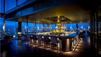 The image shows a stylish, modern bar with an illuminated central counter, surrounded by bar stools. Floor-to-ceiling windows offer a stunning night view of a cityscape, adding to the sophisticated ambiance with soft, warm lighting throughout the space.