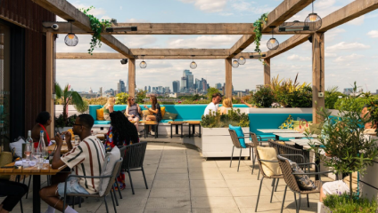 A vibrant rooftop terrace features people dining and socializing under wooden pergolas decorated with hanging lamps and plants. Modern skyscrapers form the backdrop of the city skyline, framed by a partly cloudy sky. The seating includes a mix of tables and benches.