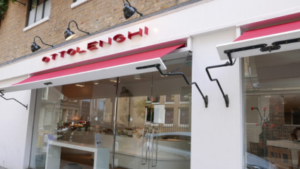 A storefront with a red and white sign reading OTTOLENGHI features large windows and modern awnings. The building has a brick facade above the shop, and street reflections are visible in the windows. Inside, there are tables, chairs, and a counter visible.