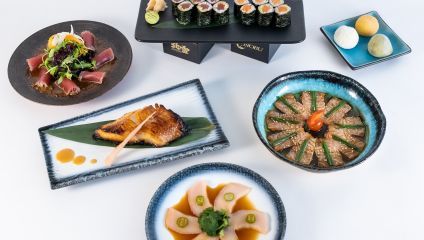 A spread of Japanese cuisine is displayed on a white surface, featuring sushi rolls, a plate of sashimi garnished with greens, a dish of grilled fish, thinly sliced fish in a blue bowl with sauce, another sashimi dish, and three spheres of mochi dessert.
