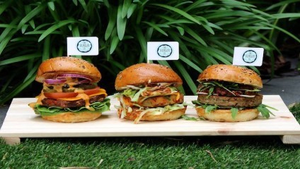 Three gourmet burgers are displayed on a wooden board against a backdrop of green leafy plants. Each burger has a small flag with a logo on top and includes a variety of ingredients like fresh lettuce, cheese, tomato, and onions in a toasted bun.