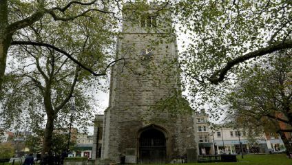 A tall stone church tower, topped with a clock showing the time as approximately 10:12, stands amidst leafless trees in a park. Below the clock, there is an arched entrance. Buildings and shops are visible in the background under a cloudy sky.