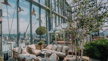 A stylish rooftop terrace with wicker furniture, plush cushions, a small white greenhouse, potted plants, and decorative metal birds suspended in flight. The space offers a panoramic view of the city skyline through tall glass walls.