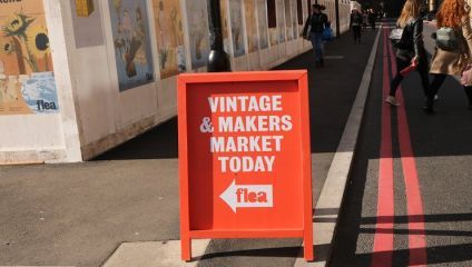 A red sign with white text that reads VINTAGE & MAKERS MARKET TODAY and an arrow pointing left towards flea stands on a city sidewalk. Several people are walking along the pathway beside some wall posters.