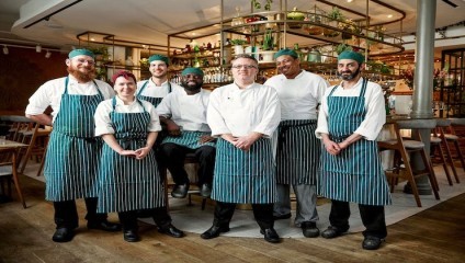 A group of six chefs stand together in a modern restaurant kitchen, all wearing striped aprons and chef hats. Smiling, they pose confidently, with various kitchen tools and plants visible in the background. The atmosphere appears bright and welcoming.