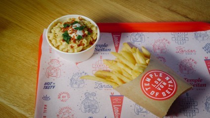 A red tray with a bowl of macaroni and cheese garnished with herbs and a dollop of sauce alongside a serving of fries in a paper holder, resting on a Temple of Seitan branded paper liner atop a wooden table.