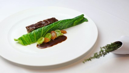 A gourmet dish is artfully presented on a white plate. The dish includes a neatly wrapped green leaf, segments of green grapes, roasted vegetables, and a brown sauce. A sprig of fresh herbs is placed beside the plate for garnish.