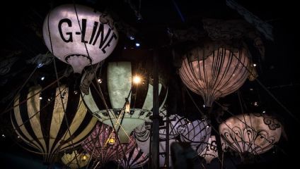 A cluster of hot air balloons with various designs and patterns are dimly lit against a dark background. The balloon in the foreground has G-LINE written on it. The scene has a whimsical and magical atmosphere.
