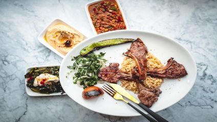 A white plate with grilled lamb chops served with rice and sautéed greens, garnished with a grilled green pepper on a marble surface. Surrounding the plate are three small dishes, one with hummus, another with a spicy sauce, and one with mixed greens and cheese.