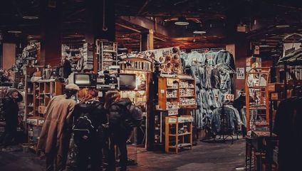 A dimly lit indoor market features various stalls. One prominent booth in the center showcases vintage electronics, including old televisions. To the right, a display of denim jackets is visible. Several people are browsing the items. The atmosphere is cozy and nostalgic.