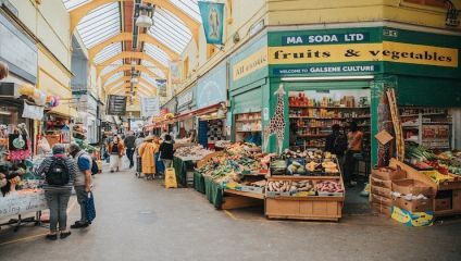 A bustling indoor market with various stalls selling fruits and vegetables. Shoppers walk along a wide aisle under a high, arched yellow ceiling with large windows. Brightly colored produce is displayed in open boxes and baskets, creating a vibrant scene.