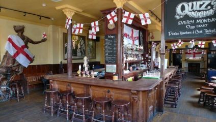 A bar with wooden decor is adorned with English flags. A chalkboard advertises a quiz night. Stools surround the bar, which is stocked with bottles and glassware. In the background, there's a large statue of a figure with a flag and a painting on the wall.