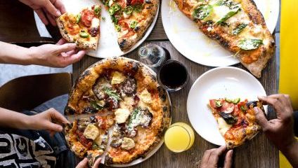 A group of people sitting around a wooden table enjoying a meal with three large, freshly-baked pizzas topped with various ingredients like cheese, pepperoni, jalapeños, and basil. A glass of orange juice and a glass of dark soda are also on the table.
