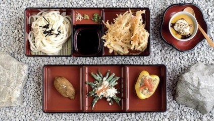 A traditional Japanese meal is served in a bento box placed on a bed of gravel. It contains udon noodles with garnish, a sesame dipping sauce, vegetable tempura, a chestnut, green beans in a sauce, and a stuffed yellow pepper. A bowl of sauce with grated radish is nearby.