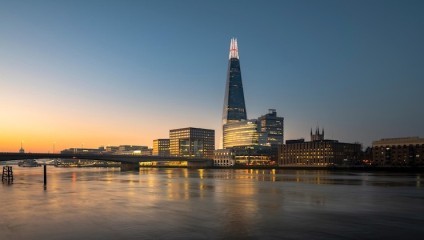 A cityscape of London at sunset featuring the Shard skyscraper prominently. The Shard is illuminated, overlooking the Thames River with reflections on the water. The surrounding buildings, bridge, and the peaceful evening sky enhance the serene scene.