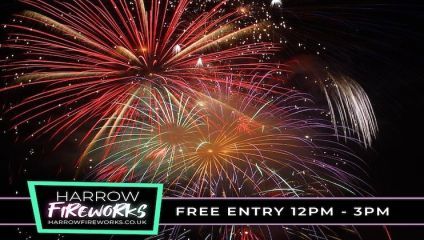 A nighttime display of colorful fireworks bursting in the sky. A sign in the bottom left corner reads Harrow Fireworks with the website harrowfireworks.co.uk, and another sign on the bottom right states Free Entry 12PM - 3PM.