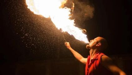 A performer with a shaved head and red sleeveless shirt breathes fire into the night sky. He holds a torch in one hand, igniting a burst of flames and creating a dramatic effect against the dark background.