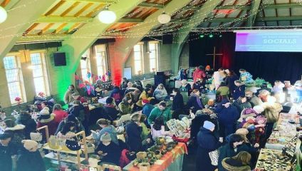 A bustling indoor market is held in a hall with wooden beams and string lights. Numerous vendors display crafts, food, and handmade goods at decorated tables. Shoppers browse the various stalls. A projected screen shows an event announcement in the background.