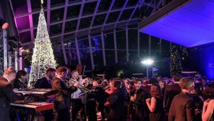 A lively indoor event features a large crowd of people dressed in formal attire, gathered under an illuminated, modern-style Christmas tree. Musicians with instruments perform on stage to the left, while colorful lights and a glass ceiling enhance the festive atmosphere.