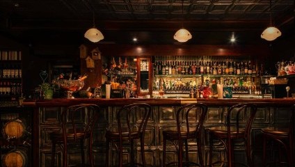 A dimly lit bar with an ornate wooden counter and high wooden stools. The bar is stocked with various bottles of alcohol and glassware. Above the counter hang three round lights, casting a warm, inviting glow. Shelves and decorations adorn the back wall.