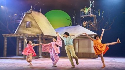 Four actors, dressed in vibrant clothing, joyfully hold hands and dance on stage. The backdrop features two wooden structures, one resembling a house and the other elevated with people inside. A large green circle is visible, creating a whimsical atmosphere.