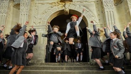 A group of children in school uniforms are joyfully jumping and cheering on the steps of a historic building with ornate carvings. Some are mid-air, displaying excitement and energy. The students are dressed in grey blazers, ties, and skirts or shorts.