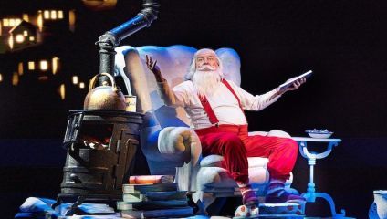 A person dressed as Santa Claus, with a white beard and red attire, sits on a large white armchair holding a book. Nearby is a vintage stove with a kettle on top, surrounded by stacked books. Christmas lights and festive decorations are visible in the background.