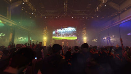 A large crowd enjoys a concert or live event in a dimly lit indoor venue, with a massive screen displaying dynamic visuals. The atmosphere is vibrant with multicolored lights, confetti, and smoke effects. People in the audience have their hands raised, capturing the moment.