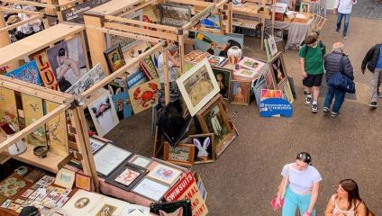 A bustling marketplace with various stalls displaying framed artwork and vintage items. Shoppers peruse the array of paintings, posters, and collectibles. Two people walk by in front, while others browse through the stalls.