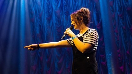 A person with their hair tied up is performing on stage, holding a microphone with one hand and pointing with the other. They are wearing a striped shirt and black overalls. The background features a lit red and blue curtain.