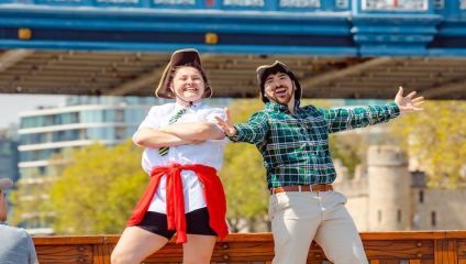 Two people wearing pirate hats stand with exaggerated poses in front of a bridge. One person, wearing a white shirt, shorts, and a red bandana around their waist, has arms crossed while smiling. The other person, in a green plaid shirt and beige pants, has arms outstretched.