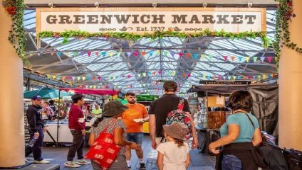 A family of four walks into Greenwich Market, a large indoor market with a canopy ceiling. Colorful bunting hangs overhead. Stalls with various items on display and people shopping are seen in the background. The market entrance sign reads GREENWICH MARKET Est. 1737.