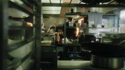 A busy restaurant kitchen with two chefs cooking at stoves amidst various pots, pans, and utensils. The foreground shows metal shelves and cooking equipment, while the background reveals the chefs focused on preparing meals under warm, dim lighting.
