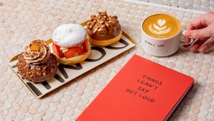 A hand holding a latte with foam art, next to a red notebook titled Things I Can't Say Out Loud. On a platter are three fancy doughnuts: one with powdered sugar and strawberries, one with a Ferrero Rocher-like topping, and one with caramel and peanuts.