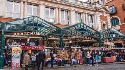 Outdoor view of Jubilee Market Hall, hosting various stalls selling goods like falafel, souvenirs, and crafts. Shoppers browse through the stalls under a glass canopy, with the market's signage visible above the entrance. A brick building and cobblestone path run along the market.