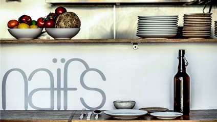 A kitchen shelf holding bowls of produce, such as tomatoes and lemons, sits above a counter. Stacks of clean plates are on the right, and utensils are placed neatly on a small plate. A glass bottle and cup are adjacent. The background features the word naïrs.