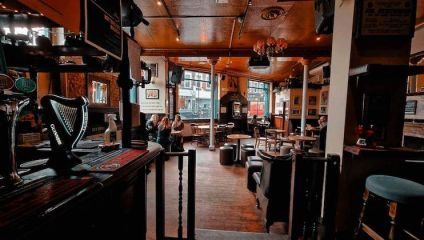 A cozy pub with wooden floors and furniture, featuring a bar with a Guinness tap in the foreground. Several patrons are sitting and chatting around tables near large windows, and natural light illuminates the space. The walls are adorned with pictures and decor.