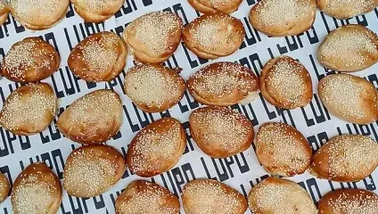 A tray covered with white paper branded LUPU holds numerous freshly baked burger buns topped with sesame seeds, arranged closely together in an even and organized manner.