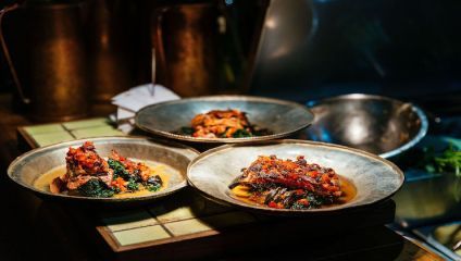 Three plates of braised meat with vegetables are displayed on a countertop. The meat appears to be topped with a red sauce and is served on a bed of greens. The background includes metallic kitchen utensils and a dimly lit setting, creating a warm ambiance.