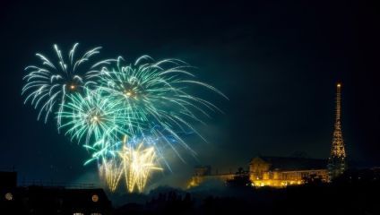 Bright green, blue, and gold fireworks burst in the night sky over an illuminated historic building and a tall, illuminated structure, creating a festive atmosphere. The dark night sky accentuates the vibrant colors of the fireworks.