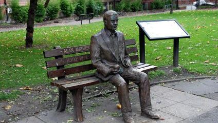 A bronze statue of Alan Mathison Turing sits on a wooden bench in a park. The plaque on the bench bears his name. The surroundings include a green lawn scattered with fallen leaves, a pathway, nearby trees, and an informational signboard.