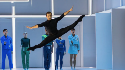 A male ballet dancer from Scottish Ballet dressed in back leaps sideways across the stage; behind him stand four dancers dressed in clinical blues and greens