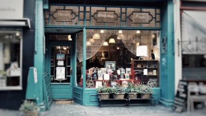 A charming bookshop with a teal storefront and gold accents displays a Bookshop, Occult, Magic sign. Through the window, various books and trinkets are visible. A few potted plants line the entrance, adding a touch of greenery to the scene.