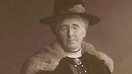 A sepia-toned photograph of an older woman wearing a dark hat adorned with feathers, glasses, and a fur-trimmed coat. She has a serious expression and is facing the camera. The background is plain and dark, emphasizing her attire and features.