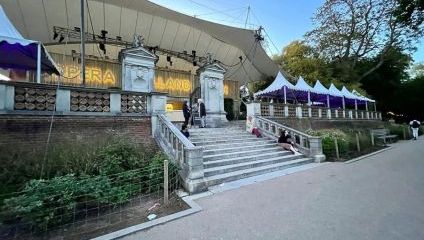 Outdoor amphitheater featuring a large stage with Opera Holland Park displayed on the backdrop. Stone steps and pillars lead up to the stage area, flanked by purple tents. Trees and a pathway surround the venue under a clear blue sky.