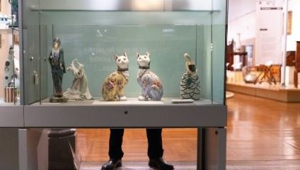 A display case in a museum contains ceramic figurines, including two cats and three human figures. One figure is a woman in an intricate dress, another a figure in a Greek-style outfit, and the third a swan. A person's legs are visible behind the case.