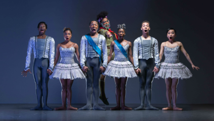 In a press shot for a production by Ballet Black, six ballet dancers stand with shocked looks on their faces whilst a seventh, behind them, reaches his arm through their flank with a menacing look on his face