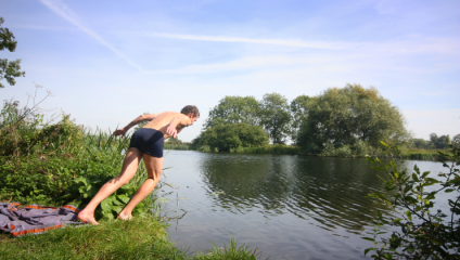 A person in swim trunks prepares to dive into a calm lake from a grassy bank. The sky is clear with a few wispy clouds, and trees line the opposite shore, reflecting on the water's surface. Nearby, a colorful towel is spread out on the grass.