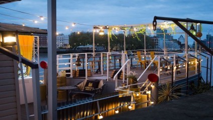 A cozy outdoor seating area on a boat at dusk, decorated with string lights and potted plants. Wooden chairs and tables are arranged on the deck. In the background, a river and some buildings are visible under a cloudy sky.