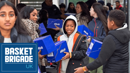 A mother and her children are amongst a crowd of people holding hampers, which will go to families struggling at Christmas, as part of charity Basket Brigade UK. The blue Basket Brigade logo is imposed in the bottom-left corner.